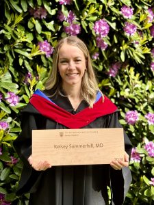 A woman in graduation attire stands in front of flowering bushes, holding a plaque that reads “Kelsey Summerhill, MD, 2024” from the University of British Columbia.